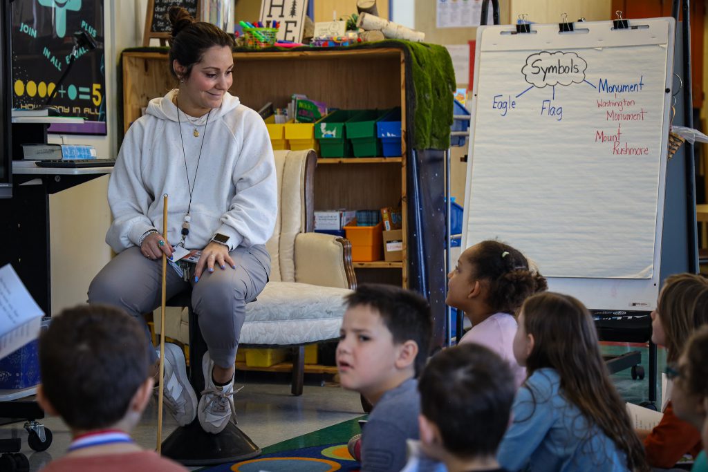 an elementary school teacher speaks to her class on the classroom carpet