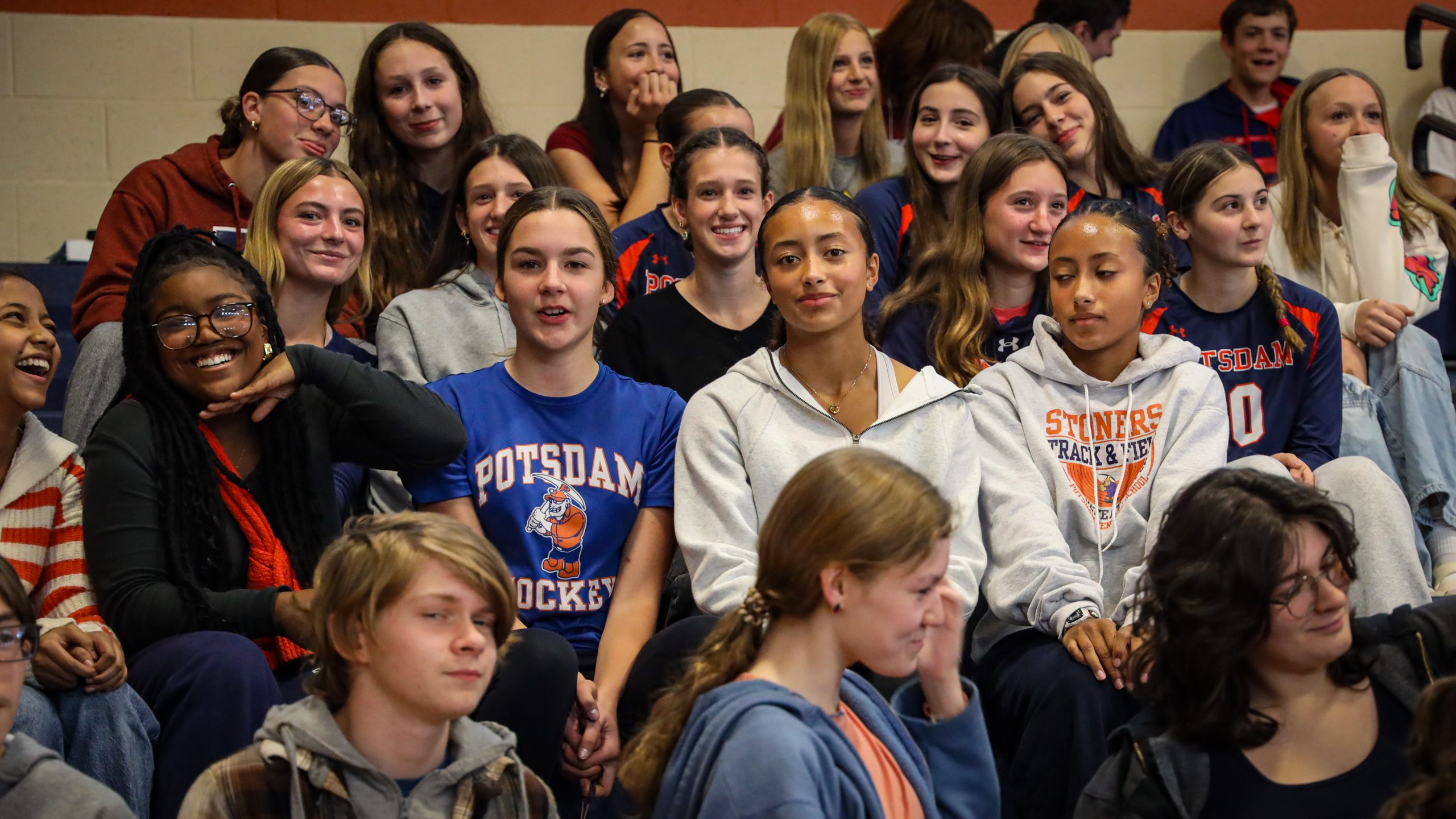 High school students sit in bleachers on pep rally day, dressed in school colors, orange and blue. They are smiling at the camera 