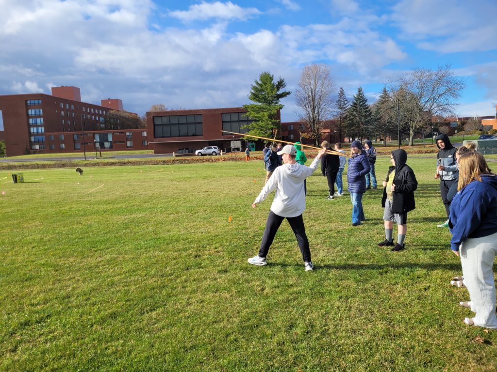 Student surrounded by peers holds large atlas dart, and prepares to throw it. 